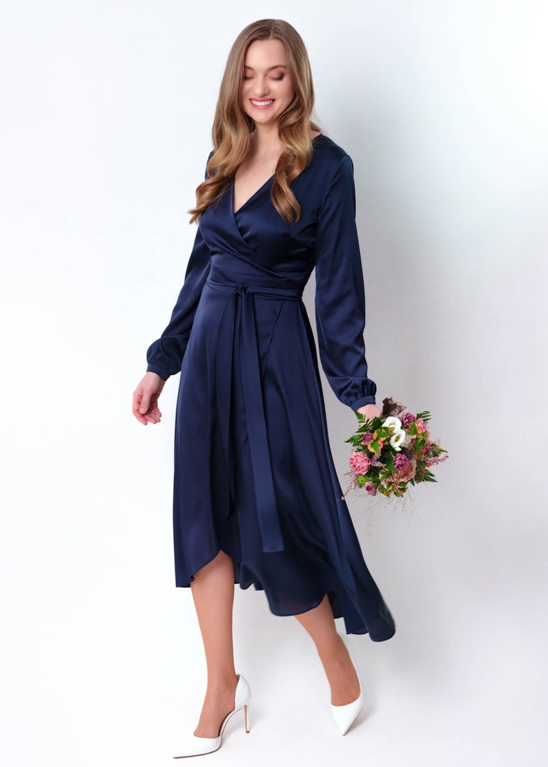 May include: A woman wearing a navy blue satin wrap dress with long sleeves and a high-low hemline. She is holding a bouquet of pink and white flowers.