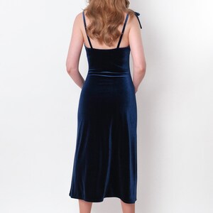 May include: A woman wearing a dark blue velvet midi dress with thin straps. The dress has a fitted bodice and a flowing skirt. She is wearing white heels.