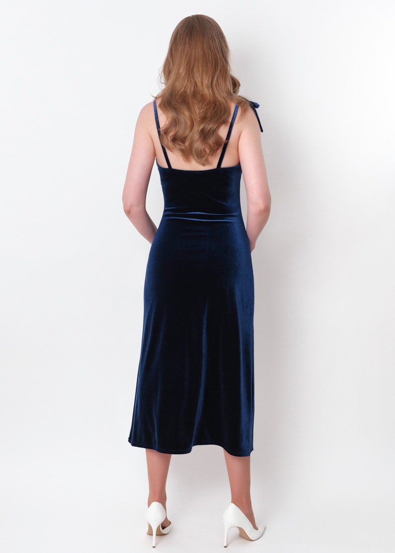 May include: A woman wearing a dark blue velvet midi dress with thin straps. The dress has a fitted bodice and a flowing skirt. She is wearing white heels.