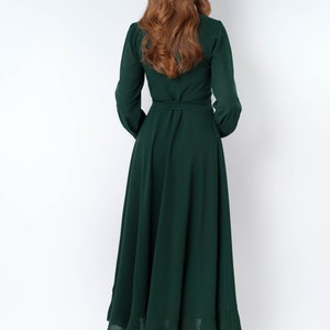 May include: A long, dark green maxi dress with a wrap style bodice and long sleeves. The dress has a ruffled hem and is cinched at the waist with a matching belt.