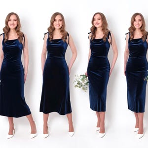 May include: Four women wearing navy blue velvet dresses with thin straps and a cowl neckline. Each woman is holding a bouquet of white flowers and is wearing white heels.