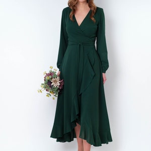 May include: A woman wearing a long-sleeved, emerald green wrap dress with a ruffled hem. She is holding a bouquet of flowers and is wearing white heels.