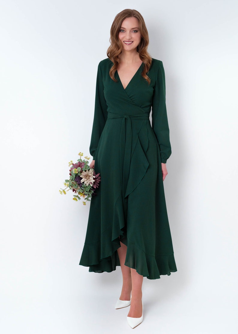 May include: A woman wearing a long-sleeved, emerald green wrap dress with a ruffled hem. She is holding a bouquet of flowers and is wearing white heels.