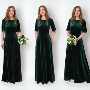 May include: Three women wearing long, emerald green velvet dresses with short sleeves and a round neckline. Each woman is holding a bouquet of white flowers.