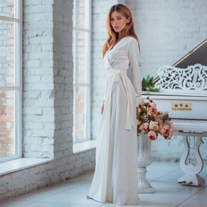 Op de afbeelding: Een lange, vloeiende witte wikkeljurk met lange mouwen en een strik in de taille. De jurk wordt gedragen in een kamer met een witte piano en een boeket roze bloemen. De achtergrond heeft een witte bakstenen muur en een groot raam.