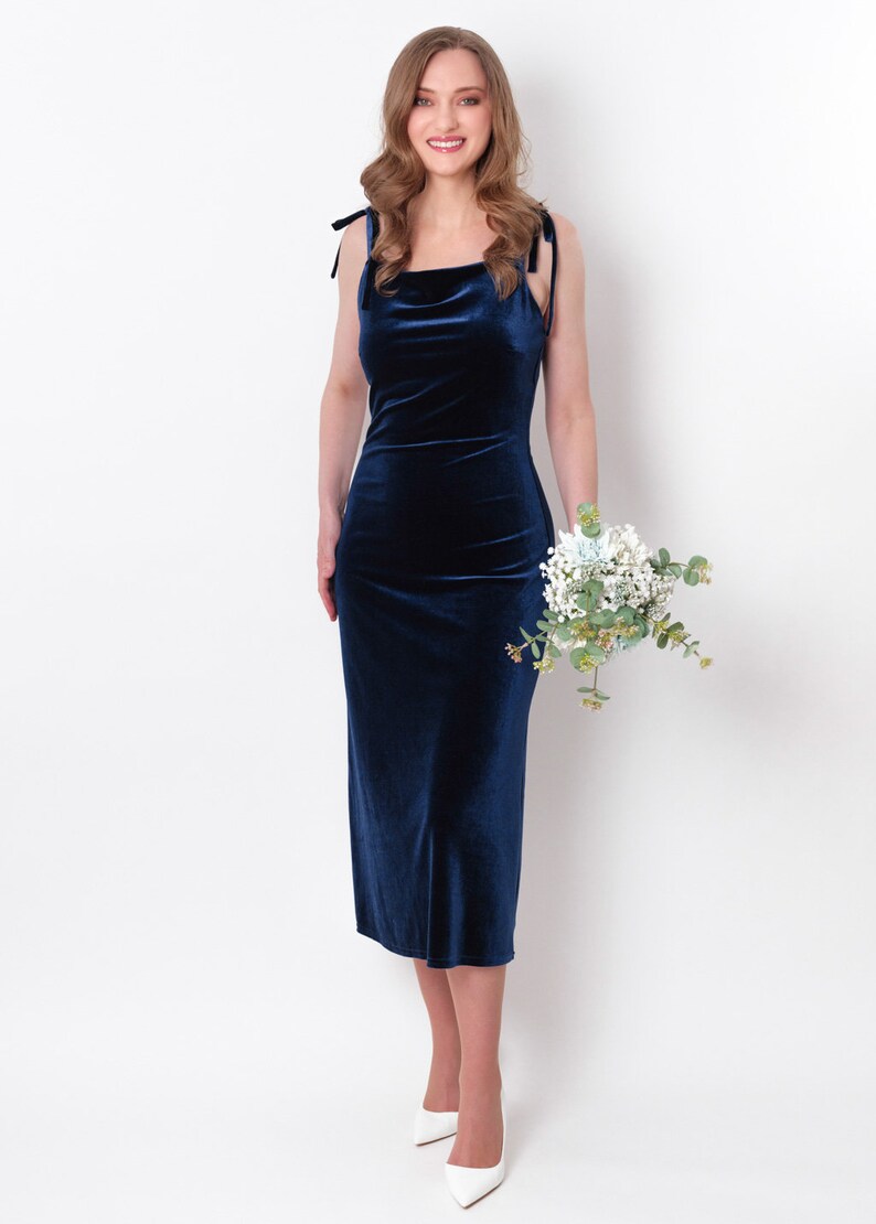 May include: A woman wearing a navy blue velvet midi dress with thin straps and a square neckline. She is holding a bouquet of white flowers and greenery.
