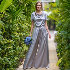 May include: A woman in a silver jumpsuit with a cowl neck and wide legs, standing on a path in a garden, holding a bouquet of blue flowers.