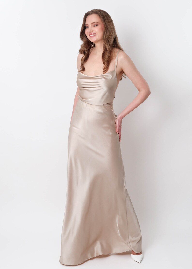 May include: A woman wearing a long, flowing, champagne-colored satin dress with a cowl neck and spaghetti straps.