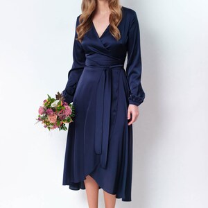 May include: A woman wearing a navy blue satin wrap dress with long sleeves and a tie at the waist. She is holding a bouquet of pink and white flowers and is smiling at the camera.
