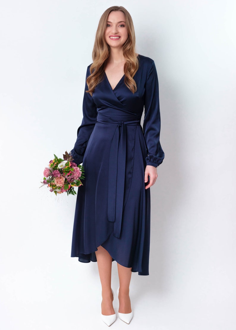 May include: A woman wearing a navy blue satin wrap dress with long sleeves and a tie at the waist. She is holding a bouquet of pink and white flowers and is smiling at the camera.
