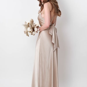 May include: A woman in a long, champagne-colored satin dress with a low back and a bow tie. She is holding a bouquet of dried flowers.