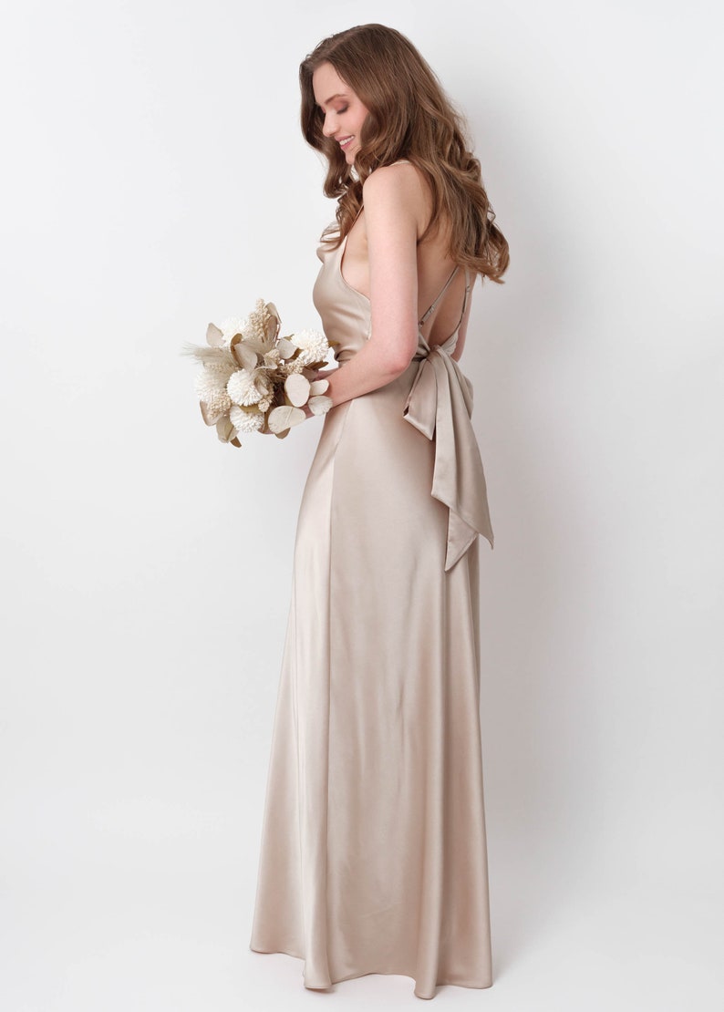 May include: A woman in a long, champagne-colored satin dress with a low back and a bow tie. She is holding a bouquet of dried flowers.
