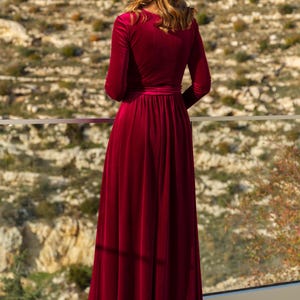K&ouml;nnte beinhalten: Ein langes, burgunderfarbenes Samtkleid mit V-Ausschnitt und langen &Auml;rmeln. Das Kleid hat ein tailliertes Oberteil und einen flie&szlig;enden Rock.