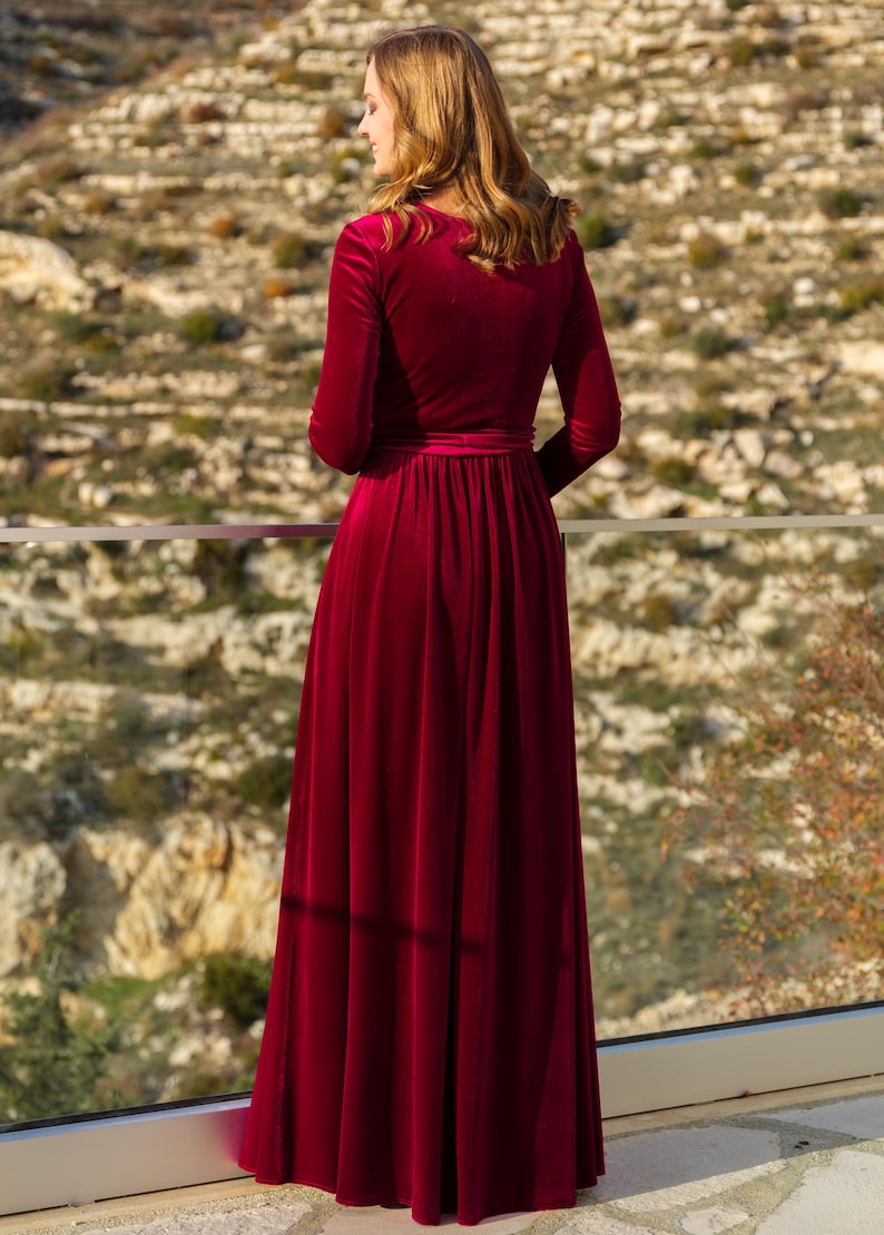 K&ouml;nnte beinhalten: Ein langes, burgunderfarbenes Samtkleid mit V-Ausschnitt und langen &Auml;rmeln. Das Kleid hat ein tailliertes Oberteil und einen flie&szlig;enden Rock.