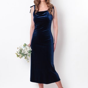 May include: A woman wearing a navy blue velvet midi dress with thin straps. She is holding a bouquet of white flowers and smiling at the camera.