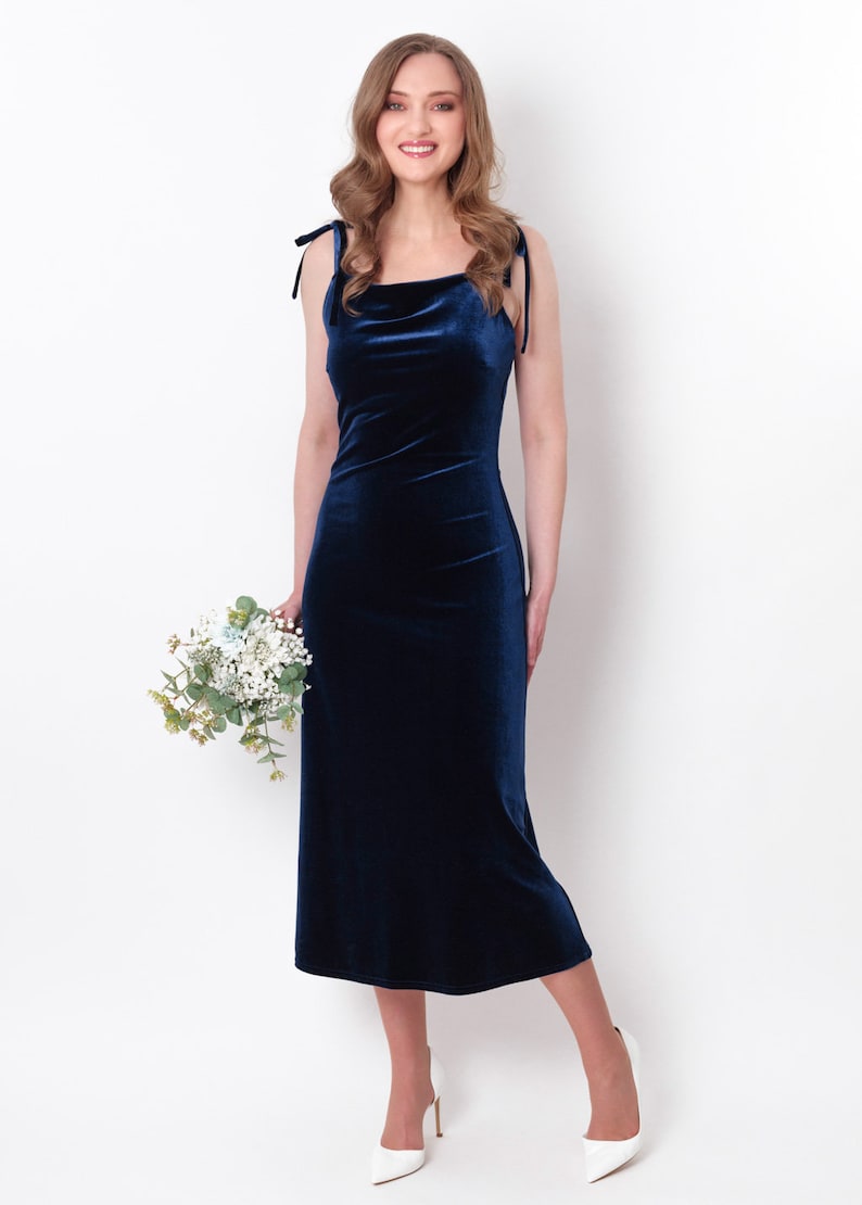 May include: A woman wearing a navy blue velvet midi dress with thin straps. She is holding a bouquet of white flowers and smiling at the camera.