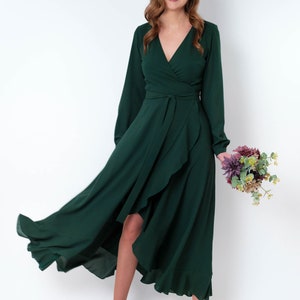 May include: A woman wearing a long, dark green wrap dress with a high-low hemline. The dress has long sleeves and a V-neckline. She is holding a bouquet of flowers.