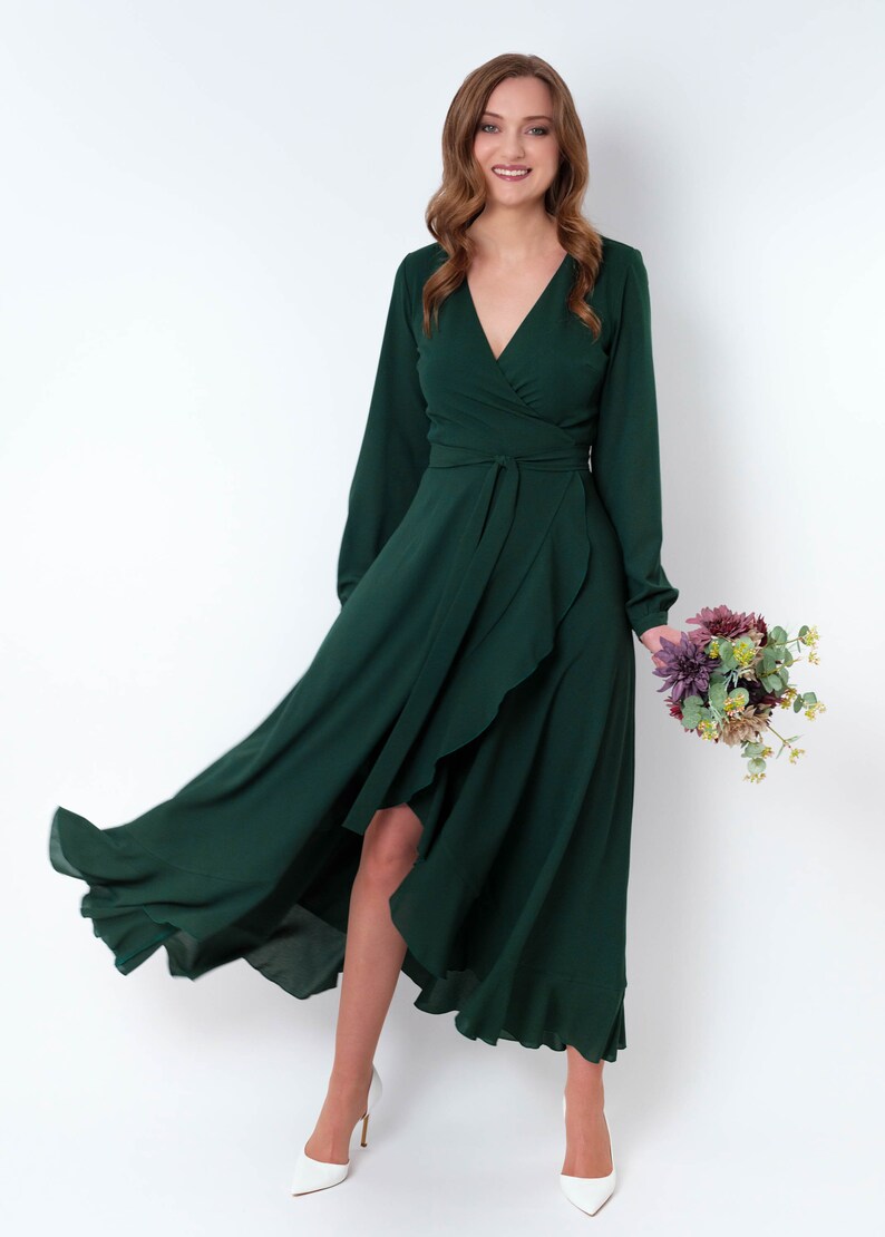 May include: A woman wearing a long, dark green wrap dress with a high-low hemline. The dress has long sleeves and a V-neckline. She is holding a bouquet of flowers.