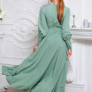 May include: A woman wearing a sage green maxi dress with long sleeves and a tie at the waist. The dress has a ruffled hem and the woman is wearing silver heels.