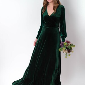 K&ouml;nnte beinhalten: Ein langes, dunkelgr&uuml;nes Samtkleid mit V-Ausschnitt und langen &Auml;rmeln. Das Kleid hat einen Wickel-Oberteil und einen flie&szlig;enden Rock. Das Model h&auml;lt einen Blumenstrau&szlig;.