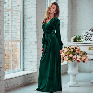 May include: A woman wearing a long, emerald green velvet dress with a V-neckline and long sleeves. She is standing in a room with white brick walls and a white grand piano in the background.