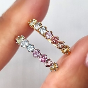 Puede incluir: Dos anillos delicados con piedras preciosas de colores. Los anillos están apilados en un dedo y presentan un degradado de piedras pastel rosa, azul y amarillo.