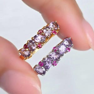 May include: Two gold rings, each featuring a row of oval purple gemstones and small red gemstones. The rings are held between fingers against a white backdrop. These are jewellery items.