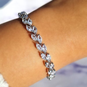 Puede incluir: Una pulsera plateada con un diseño en forma de hojas. Cada hoja está compuesta por múltiples cristales claros y brillantes. La pulsera se muestra en una muñeca, resaltando su apariencia delicada y elegante. Los cristales capturan la luz, creando un efecto deslumbrante.