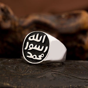 Può includere: Un anello d'argento con una pietra ovale nera con calligrafia araba. L'iscrizione recita "Allah" in lettere bianche.