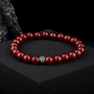 May include: A red carnelian gemstone bracelet with a silver bead accent. The bracelet is made of round beads and is approximately 8 inches in length.