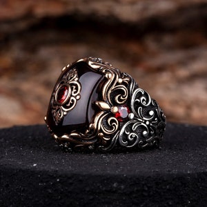 Puede incluir: Un anillo de plata con una gran piedra preciosa roja oscura y detalles intrincados y ornamentados de plata. El anillo tiene piedras preciosas rojas más pequeñas a los lados.