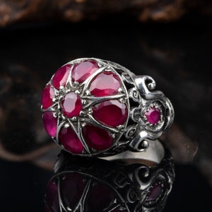 Könnte beinhalten: Ein silberner Ring mit einem großen, roten Edelstein, der von kleineren roten Edelsteinen umgeben ist. Der Ring hat ein filigranes Design mit einem floralen Motiv.