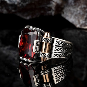 Könnte beinhalten: Ein silberner Ring mit einem großen roten Edelstein und einem goldenen Geschossdetail. Der Ring hat aufwendige silberne Designs auf der Band.