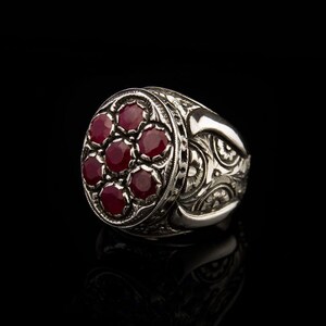 Può includere: Un anello d'argento con una montatura rotonda di rubino rosso. L'anello presenta intricati disegni floreali incisi sulla fascia.
