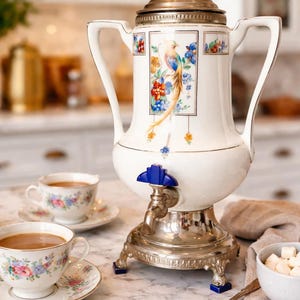 May include: A vintage white porcelain coffee urn with a bird and floral design, silver accents, and a blue spigot. Two floral teacups and saucers sit nearby, along with a bowl of sugar cubes and a spoon. The urn is approximately 18 inches tall.
