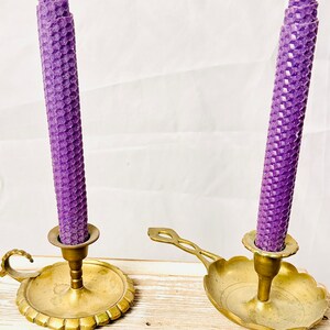May include: Two purple honeycomb candles in brass candle holders. The candle holders have a decorative design and are placed on a wooden surface.