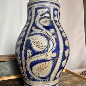 May include: A tall, antique stoneware pitcher with a blue and white floral design. The pitcher features a dark metal lid and a detailed pattern of leaves and vines. The ceramic vessel has a bulbous shape and is likely used for serving beverages.