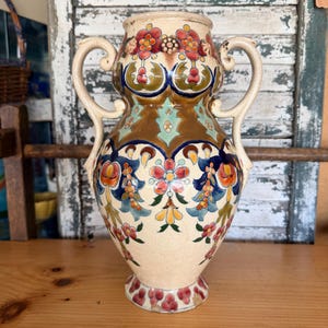 May include: A cream-colored ceramic vase featuring two curved handles and a decorative pattern of red, orange, yellow, and blue floral motifs. The surface has a visible crackle glaze finish with metallic gold accents around the neck and body.