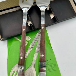 May include: A vintage salad fork and spoon set. The stainless steel utensils have dark brown wooden handles with a silver rivet. The set is displayed on a green paper with text and a black and gold box.