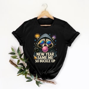 Op de afbeelding: Zwart T-shirt met een wasbeer met een zonnebril en een hoodie, die een kauwgomballenblaas blaast. De tekst "NEW YEAR SAME ME. SO BUCKLE UP" staat eronder. Kleurrijk vuurwerk is op de achtergrond.