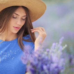 May include: A woman wearing a blue t-shirt with a white dandelion design and a straw hat, standing in a field of lavender.