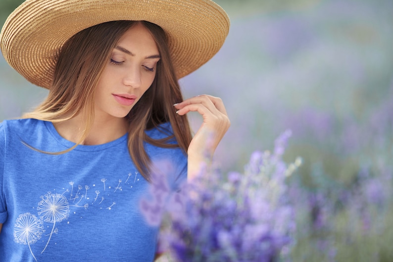 May include: A woman wearing a blue t-shirt with a white dandelion design and a straw hat, standing in a field of lavender.