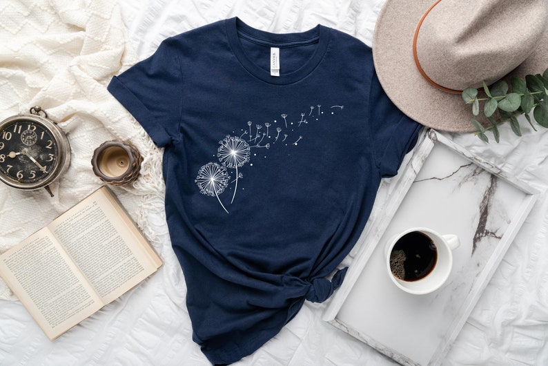 May include: A navy blue t-shirt with a white dandelion design. The dandelion seeds are blowing in the wind.