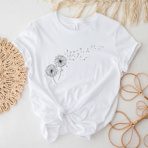 May include: White t-shirt with a black dandelion graphic. The dandelion is blowing seeds in the wind.