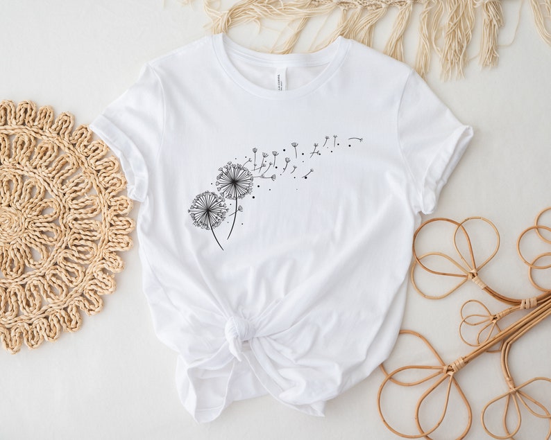 May include: White t-shirt with a black dandelion graphic. The dandelion is blowing seeds in the wind.