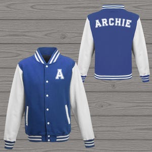 Op de afbeelding: Blauw en witte varsity-jas met de naam "ARCHIE" op de rug en de letter "A" op de voorkant. De jas heeft witte mouwen, een witte kraag en witte strepen op de manchetten en zoom. De jas wordt van voren en van achteren getoond.