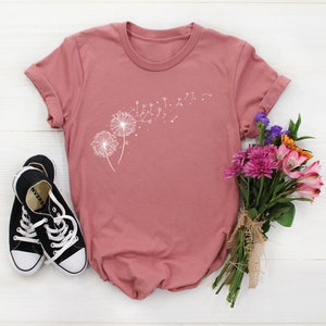 May include: A dusty rose t-shirt with a white dandelion graphic. The dandelion has seeds blowing in the wind. The t-shirt is laying on a white wooden surface with a bouquet of flowers and a pair of black sneakers.