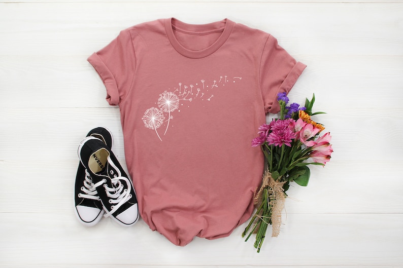 May include: A dusty rose t-shirt with a white dandelion graphic. The dandelion has seeds blowing in the wind. The t-shirt is laying on a white wooden surface with a bouquet of flowers and a pair of black sneakers.