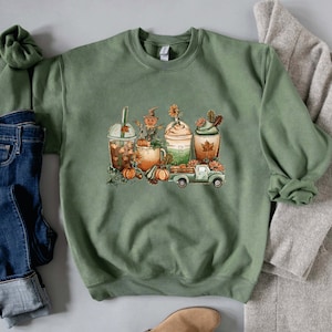 Op de afbeelding: Een groen sweatshirt met een afbeelding van herfstachtige drankjes, een vogelverschrikker, pompoenen en een pick-up truck. De drankjes zijn een pumpkin spice latte, een caramel macchiato en een peppermint mocha.