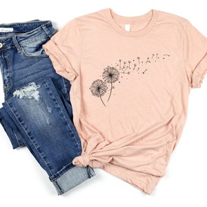 May include: A peach-colored t-shirt with a black dandelion graphic. The dandelion is blowing in the wind. The t-shirt is paired with blue jeans.